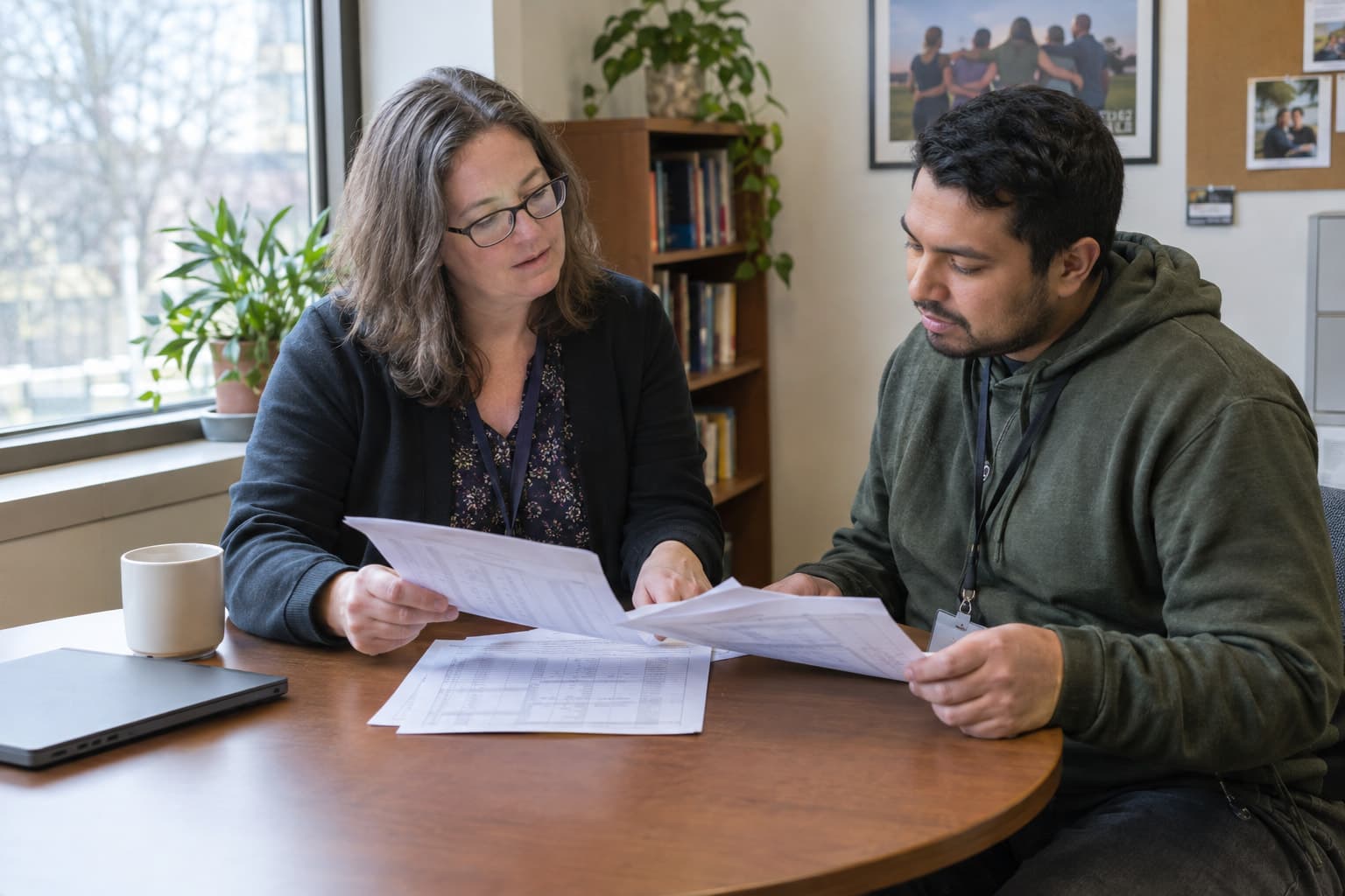Two people reviewing printed records together at a small table by a window