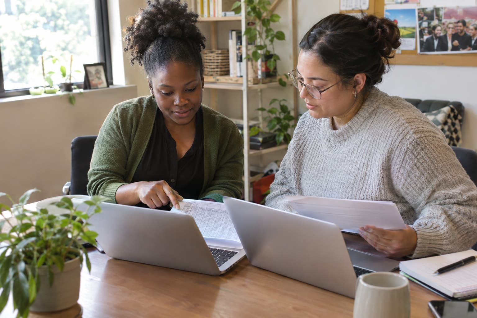 Two staff members comparing printed sheets against a laptop at a shared desk