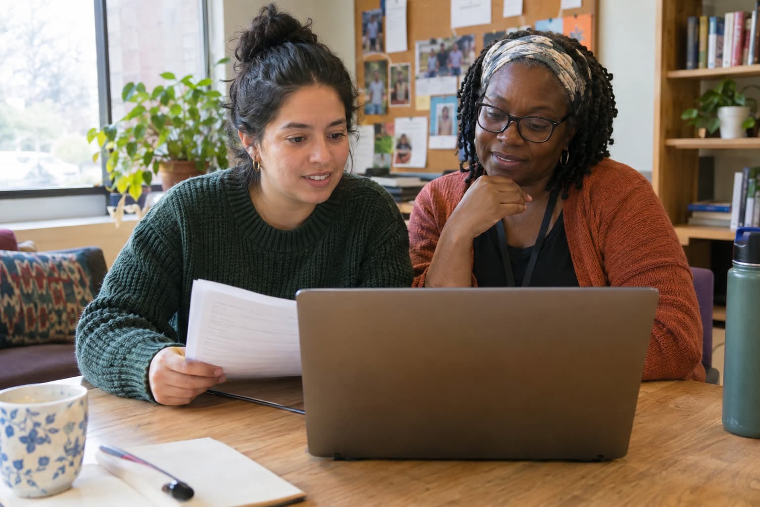 Two staff members at a shared table looking at a laptop, one holding a printed page