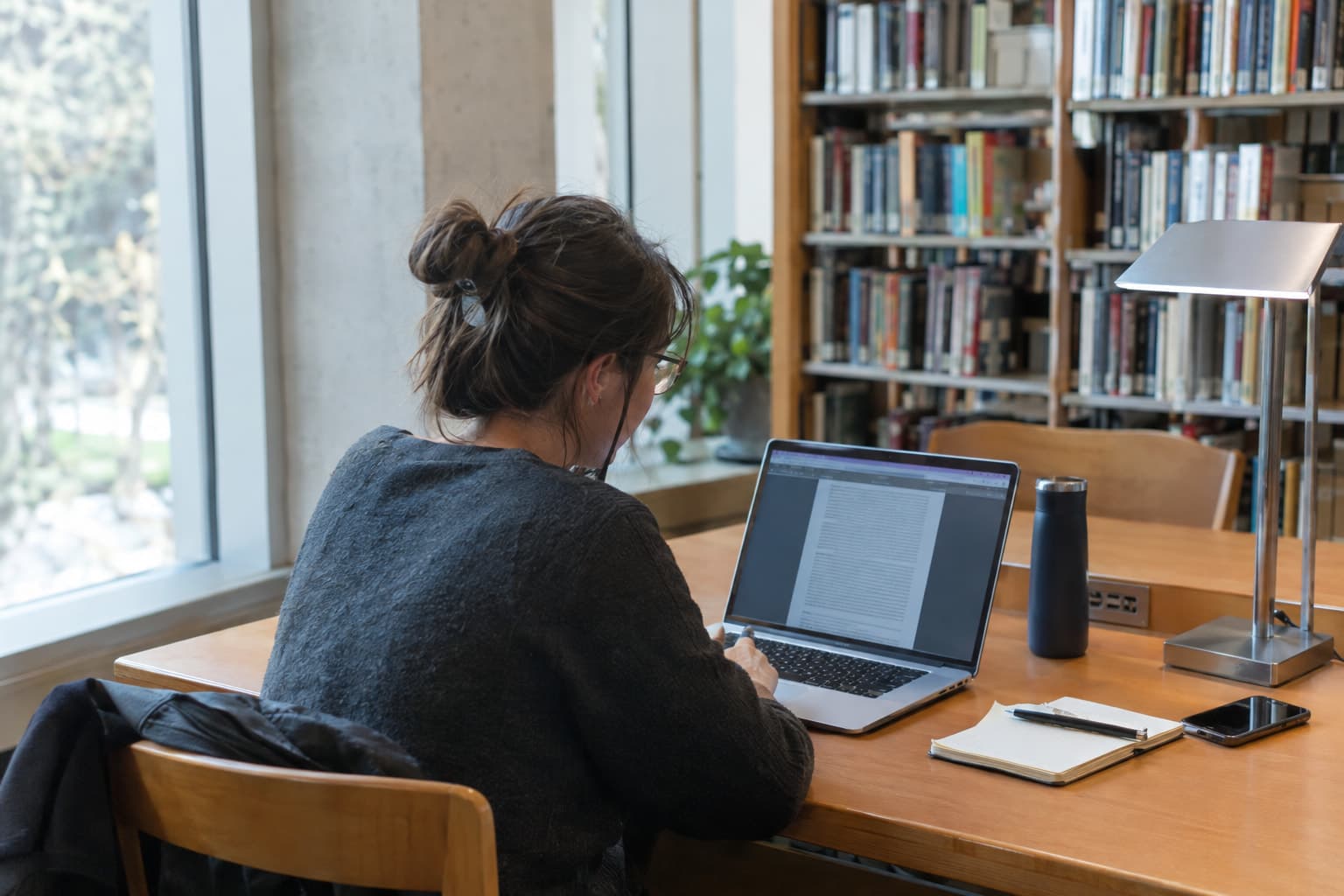 A person at a library desk reading a document on a laptop by a side window