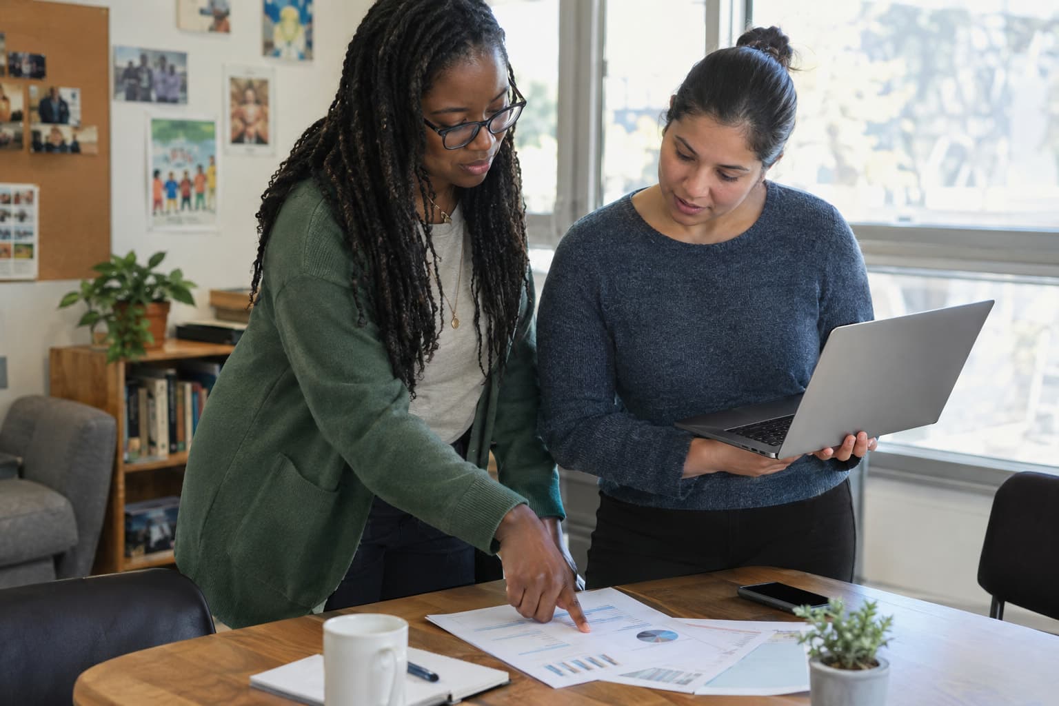 Two colleagues at a shared table, one pointing at a printed page while the other holds a laptop