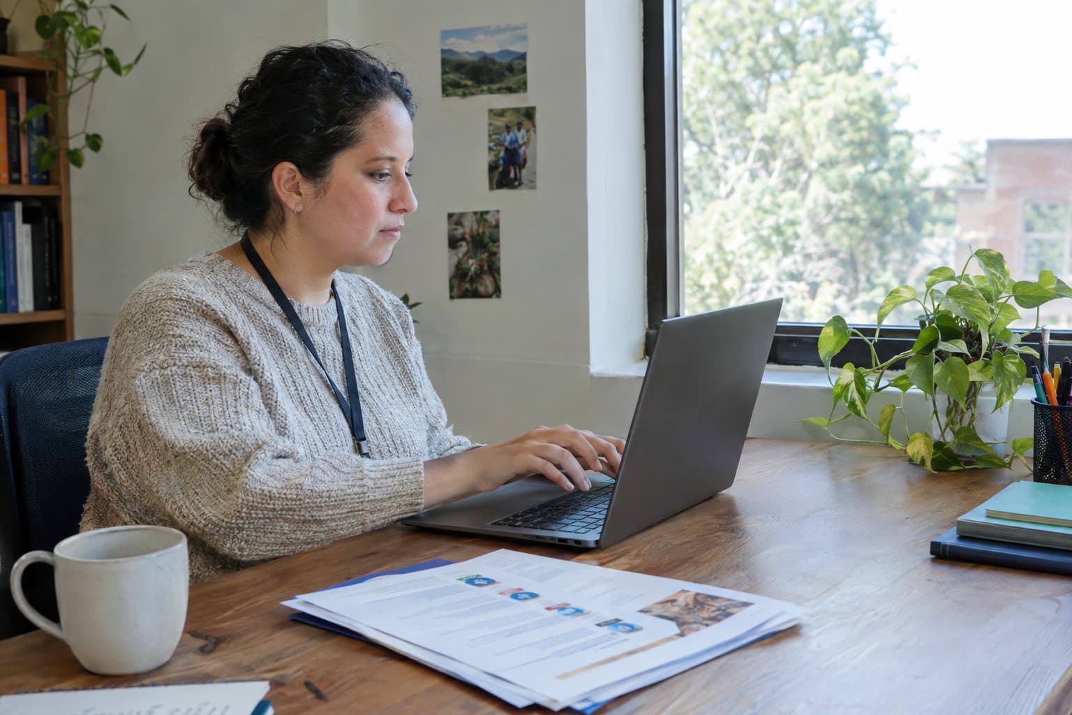 A staff member typing at a laptop near a window with printed pages on the desk