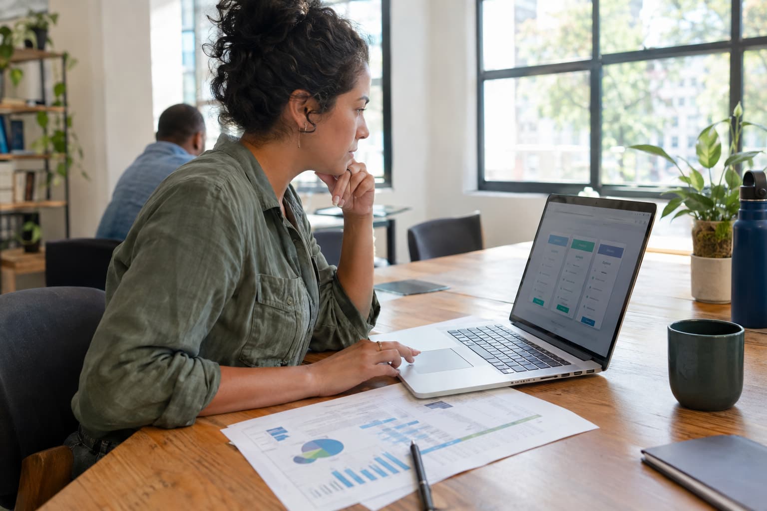 A staff member at a shared table with a laptop and printed budget sheets