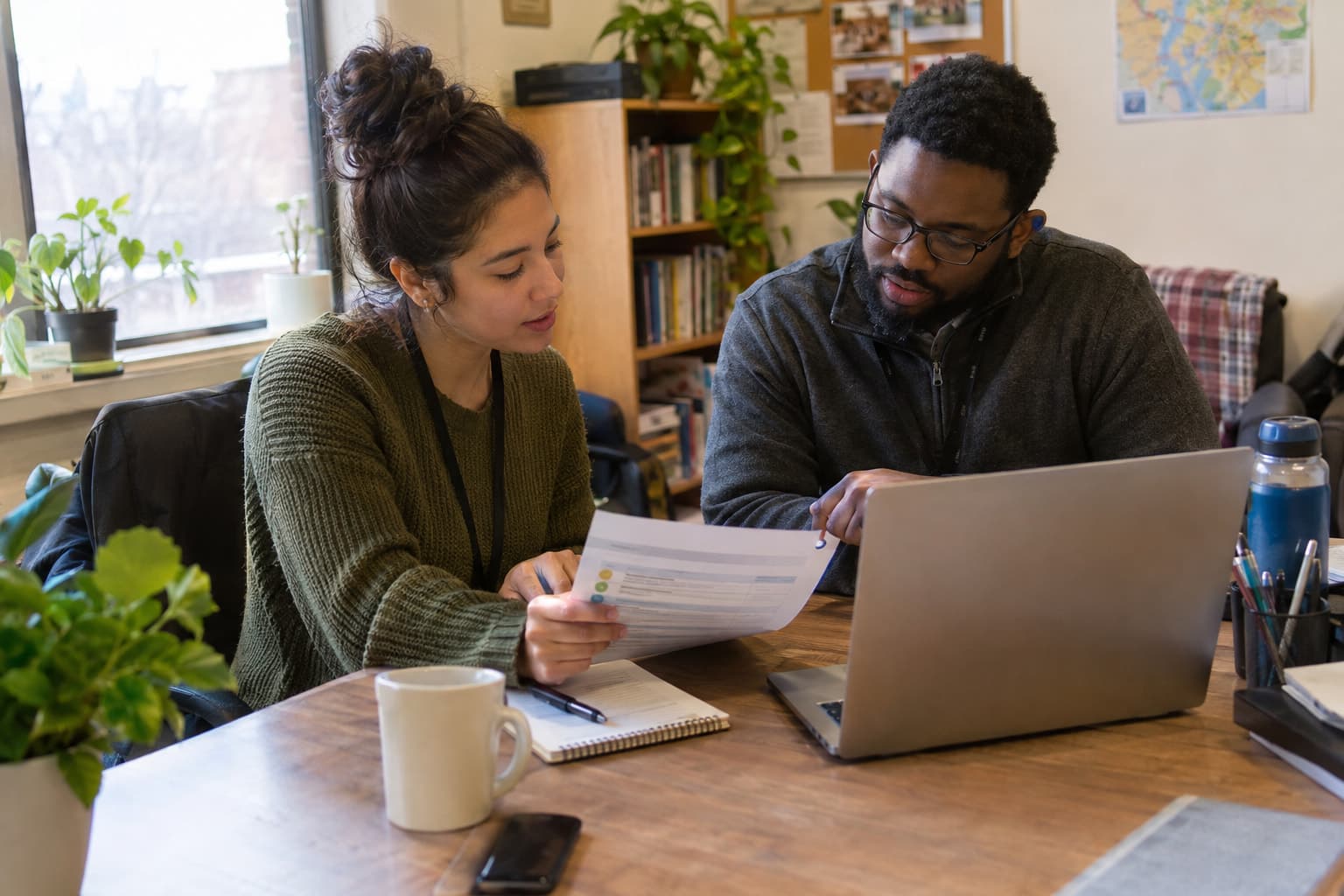Two staff members at a shared desk comparing a printed checklist with a laptop screen