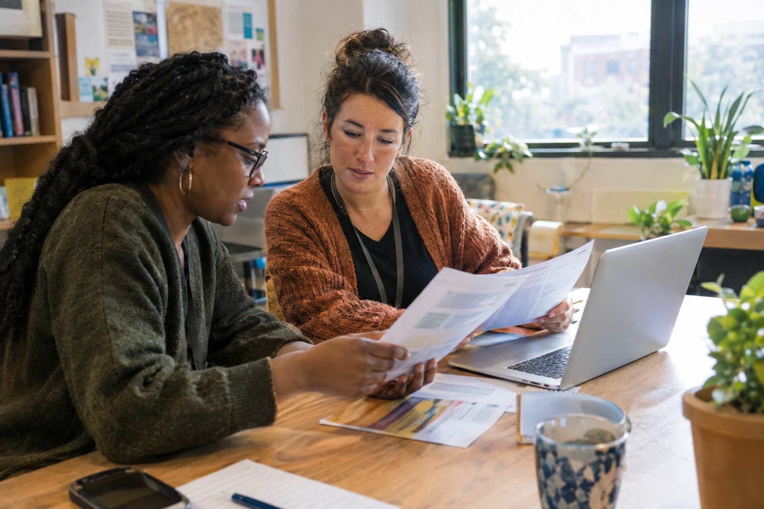 Two staff members at a shared desk comparing printed sheets against a laptop