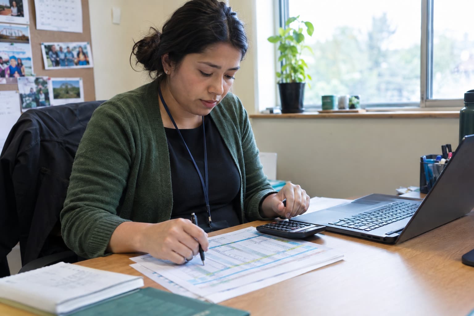 A staff member at a desk with a printed table and a calculator beside a laptop