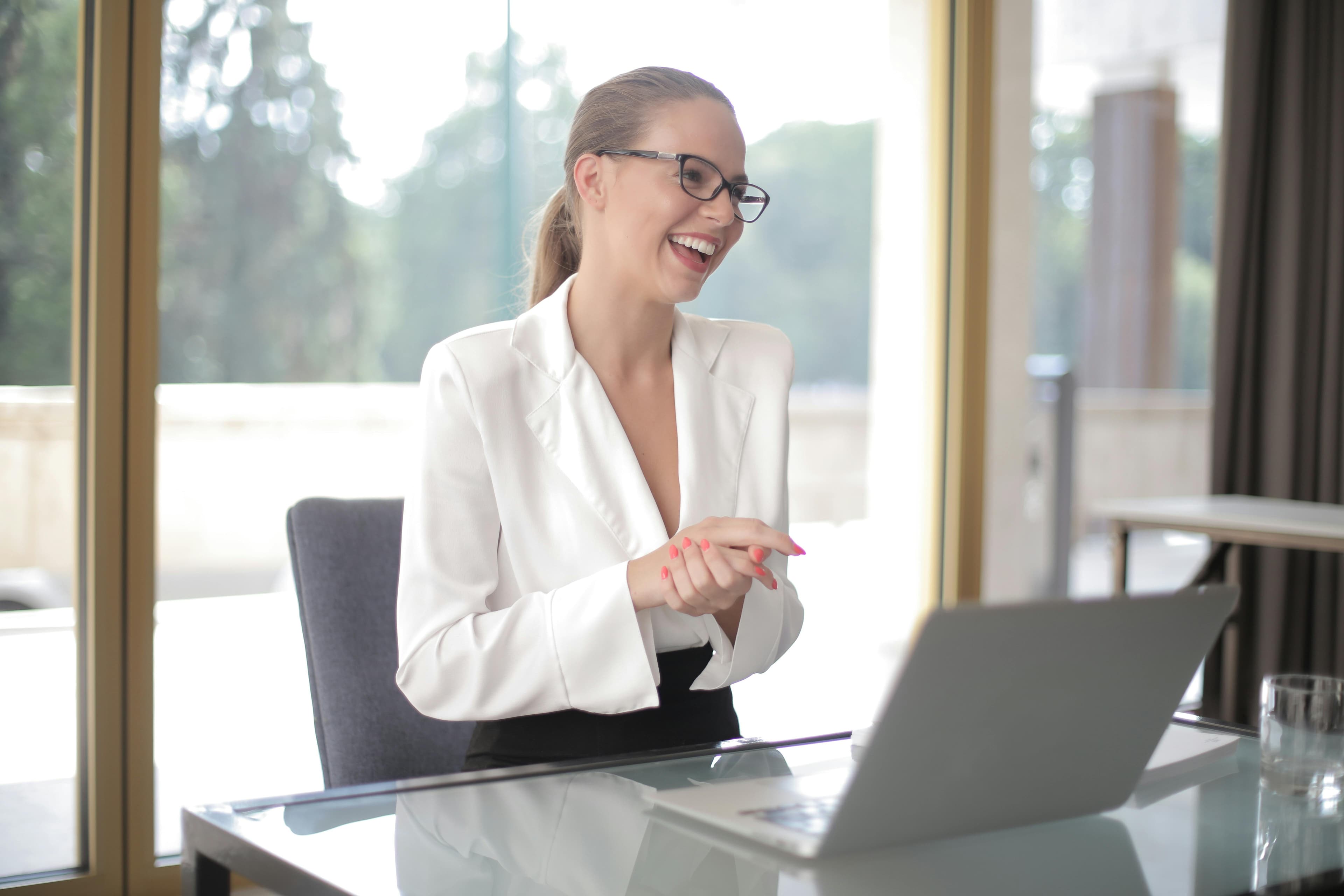 Smiling professional in a white blazer at a glass desk with a laptop, beside tall windows
